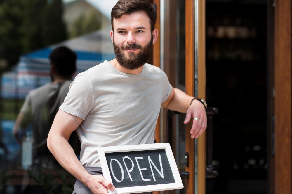 Homem segurando uma placa de "Open" na entrada de um estabelecimento, representando a formalização de um negócio. Saiba como abrir um MEI e regularizar sua empresa.
